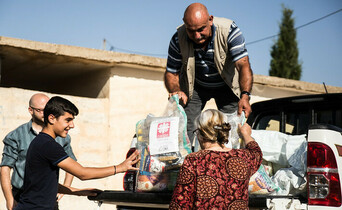 Ein Mann steht auf der Ladefläche eines kleinen LKWs und gibt Säcke mit Gewand an Menschen unten weiter. Auf den Säcken ist das Logo von Caritas Syrien.