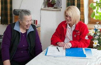 Eine ältere Frau sitzt mit einer Caritas-Mitarbeiterin am Tisch. Sie lachen.