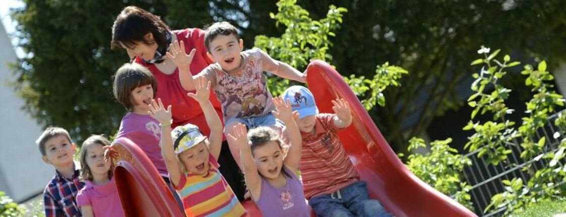 Eine Gruppe von kleinen Kindern rutscht gemeinsam eine breite Rutsche hinunter. Dahinter stehe eine Aufsichtsperson.