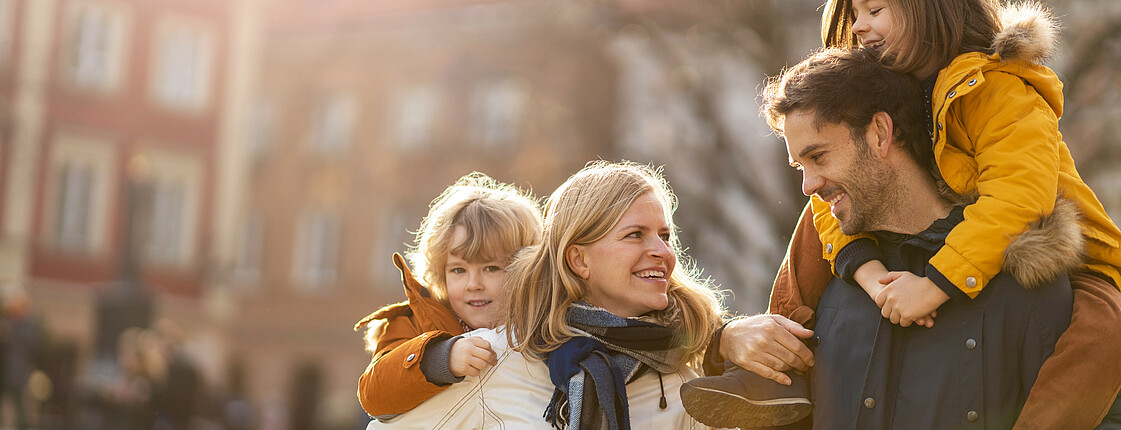 Eine gut gelaunte Familie in Winterkleidung geht bei Sonnenschein durch die Stadt. Vater und Mutter tragen jeweils ein Kind auf den Rücken.