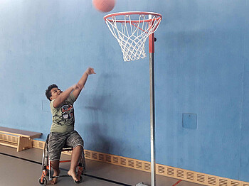 ein Bub sitzt in einem Rollstuhl und wirft einen Ball in den Basketballkorb